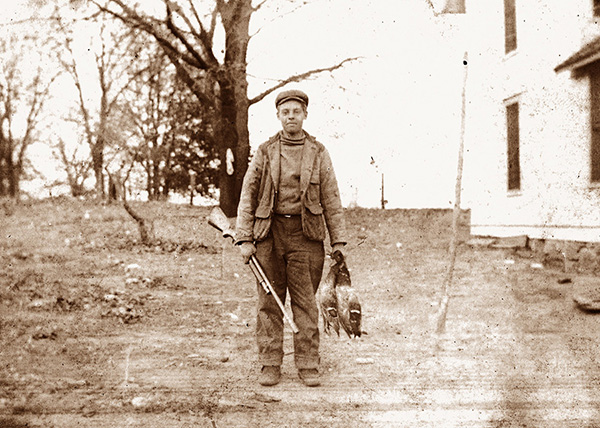 Sepia-toned photograph, likely dating to the 1930s, of a man named Harry Kenyon. Mr. Kenyon is dressed in thick hunting clothes with a billed cap. He is holding a long gun in his right hand and a pair of dead Mallards in his left hand. The outdoor photo was snapped in a large cleared space that is likely a yard or field, with a few winter-bare trees in the background and a two-story white building partially visible at right of frame.