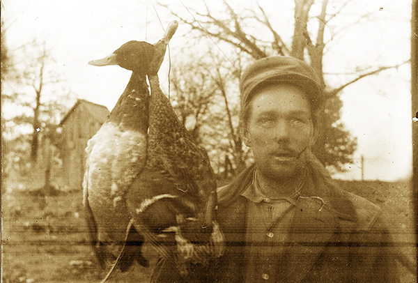 Blurry sepia-toned photo of a man standing beside two dead Mallards, which are hanging from a string or wire so that they are at head-height. The man is wearing a brimmed hat, a buttoned-up shirt with a rumpled collar, and a padded coat that appears to be made of canvas or similar material. The Mallards are male and female, judging by their plumage, and one of the female's wings hangs in way that suggests the wing was broken when she was shot. The outdoor scene shows trees that have lost their leaves for winter and a very small wooden outbuilding with a tin roof. The outbuilding looks to me like an outhouse or smoke shed.