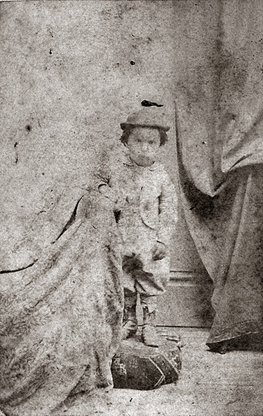 Grainy and faded grayscale portrait of a toddler. The child is standing on an upholstered footrest and is wearing knee breeches, a short jacket with long sleeves, a rounded hat with a small rolled brim, and rumpled boots. One arm rests on a curtain-draped prop. Another heavy drape hangs in the background. Time has robbed the image of most facial features, leaving only the child's dark eyes staring into the lens.