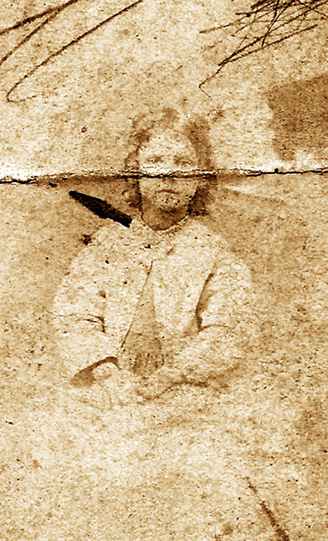 A faded, folded, and foxed photo from February 1, 1887. The child's image has almost completely faded. Loose curly hair, a long-sleeved jacket buttoned at the neck, and what might be a pleated waistline for a dress. Hands folded in lap. Serious eyes peer out over the photo's broken fold.