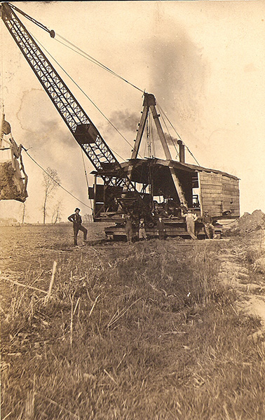 Sepia-toned photograph of a steam crane and six workers in a field in Iowa, c. 1900. The field has been cut to stubble. The steam crane is belching smoke, and the crane's body looks like a wooden building on a massive wood platform. Mounds of bare dirt are visible in the background. The workers are lined up in front of the crane, posing for the photo. A few bare trees are on the horizon, suggesting a winter setting.