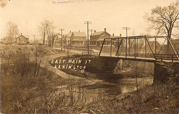 Postcard photograph stamped "Photographed by H. J. Linton" and hand-labelled "East Main St., Lexington, O". A small, single-section truss bridge crosses a brook, several two-story houses line the road beyond the bridge, and utility poles break the horizon.