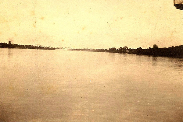 Sepia-toned image of a large body of water with a distant silhouette of truss bridge. The photo's horizon is slightly tilted, the bridge has six visible segments, and a jointed corner of wood is visible in the upper right corner of the frame.