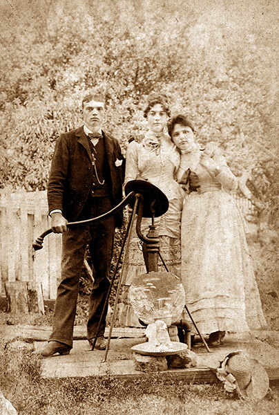 Sepia-toned photograph of three young adults posing beside a pump-handled well in what appears to be a fenced yard. All are wearing formal-appearing attire—two wearing layered and ruffled dresses and one wearing a suit with a prominent pocket-watch chain. A beribboned hat is on the ground in front of the well.