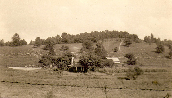 Sepia-toned photo of a house and barn at the foot of a hill, surrounded by a large empty field. The house is set within a cluster of trees, while the field has been cleared for (probably) pasture usage. A line of large rocks and boulders runs along the hill's lower slope, and a thick tree line obscures the top of the hill. A utility pole and utility lines run through the field in front of the house, and a single strand of barbed wire stretches between the camera and the farm. 