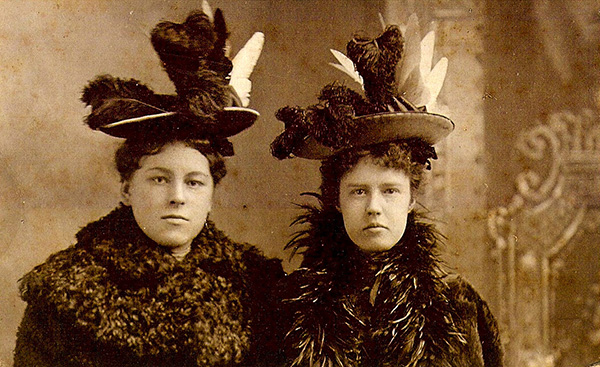 Sepia-toned portrait photograph of two women wearing elaborately decorated hats. The beribboned hats are topped with a variety of tall feathers. One woman is wearing a large fur collar, the other a flashy feather collar. Neither are smiling.