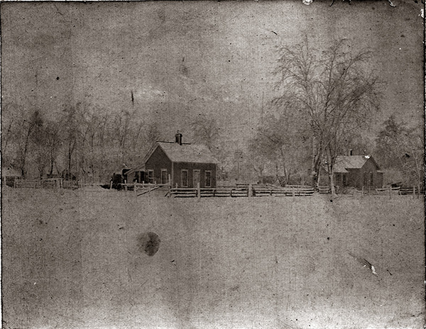 A grainy, blurred, and very faded black-and-white image of a pair of houses in a snowy setting. The houses are situation far back from the camera across a snowy field. They are encircled by a split-rail fence and winter-bare trees with snow-laden branches. The closest house appears to be somewhere between two and four rooms, with a prominent chimney in the center of the steeply peaked roof. The background house looks to have more rooms, with perhaps an addition-style room on one side. With written histories back to the 1860s, we know that the family lived as a multi-generational unit: a Civil War veteran patriarch (volunteer Ohio cavalry), his son and daughter-in-law, and their eventual six children (the youngest of which was my grandmother).