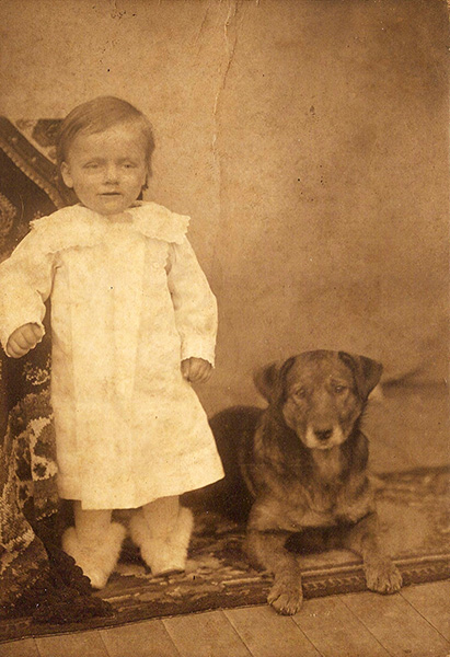 Sepia-toned photograph of a toddler in a delicately stitched white dress, standing next to an alert, mid-sized dog. The toddler's short hair is combed to one side, and their white-stockinged feet are clad in very fluffy white-furred shoes. A thick-woven rug or tapestry is draped over a support, which the toddler is leaning against, and runs across the floor. The dog is lying down, head up and looking at the camera. The dog has button ears and short-ish legs, a thick medium-length coat, and enough gray around the muzzle to suggest aging. I would guess the dog's lineage to be a terrier-type mixed with a herding-type.