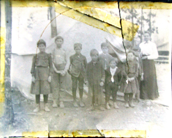 A faded black-and-white photograph of a group of children gathered in an outdoor setting front of what appears to be a tent. A chaperone in a long skirt and pale blouse stands with the children. The eight children are wearing clothes of various functional styles, including knee-length skirts, pants of full-length and knee-length, and shirts with full-length sleeves. Many are wearing coats and all are wearing work-style boots. One child, in the front row, has a more formal-appearing outfit, including a white shirt with dark pants and a dark coat fastened at a single point across the torso. The bare ground and thin evergreen trees (background), with a smudge of what appears to be winter-bare trees in the farthest background, suggests cold weather. The photo is torn in places, with yellow-tinged tape marks across the tears and along the edges.