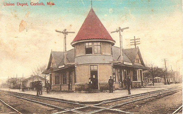 Tinted photo postcard, c. approximately 1900. Text on the image reads "Union Depot, Corinth, Miss." The image shows a train depot situated at intersecting tracks. The two-story depot features a round turret-like construction on the front, and the tile roof of the turret is tinted red. Two tall poles on either side of the turret support wires from power poles that line the streets to either side of the intersection. Several men are standing in front of the depot, and houses are visible along the streets.