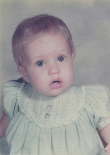 Pale-tinted photo of me somewhere in my age-counted-as-months era. In this studio-type photo, I'm gazing vaguely in the direction of the camera, wide-eyed and open-mouthed and, apparently, confused. Mother had dressed me in what was likely a stiff and itchy blue shirt with puffed sleeves and ruffles and tucks and lace (eww).