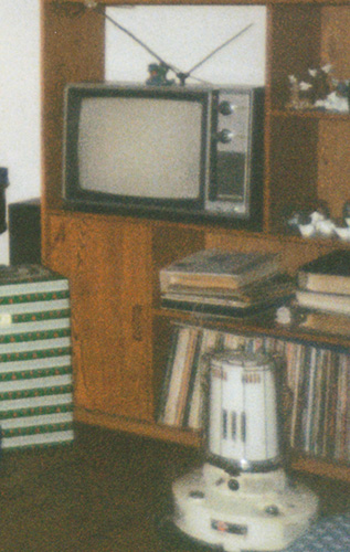 Photo of a large living-room entertainment center loaded with Christmas decorations, a console-type TV with rabbit-ears antenna, photo albums, and vinyl records. A kerosene heater, unlit, is on the floor in front of the stand, and a large wrapped gift is visible to one side.