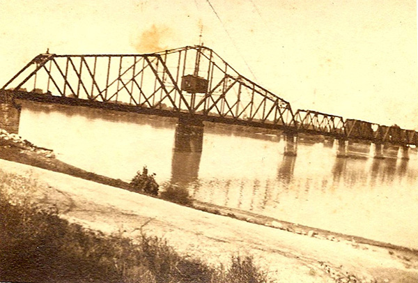 Sepia-toned image of a long truss bridge over a smooth body of water. The bridge has at least five sections set on stone pilings. A large box-like feature hangs over the bridge's final section, secured within the braces of the tallest truss-element. A bare path/lane runs across the foreground, and a treeline is just visible in the far background.