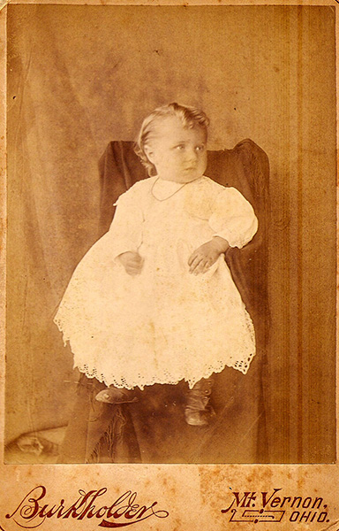 Sepia-toned studio-style photo of an very young (perhaps early toddler-aged) child seated on a draped chair. The child's blond hair is styled in short ringlets. Button-up boots and a lacy white dress, along with a dainty necklace, complete the look. The child is looking to one side with an unsmiling expression.