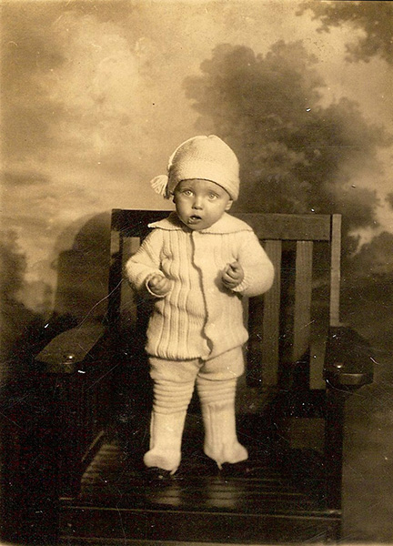 Sepia-toned studio-style photograph of a toddler. The child is dressed in an elaborately knitted outfit (perhaps an early version of the onesie?), complete with a tasseled hat and shoe-cover leggings. They are standing in a wooden chair posed against a cloudy background. I suspect there is a treat clutched in one or both hands, as the photo appears to have been snapped mid-snack. The child is peering just above and to one side of the camera with an expression that strikes me as mild confusion mixed with a touch of reluctant obedience. As if someone has told them to "stand right there and don't move a muscle" (as I was often instructed, for school photos), but the instructions were confusing because standing is chairs is typically discouraged and how can you stand still without moving a muscle but also enjoy the treat you've been bribed with? (I obviously read too much into these photos...)