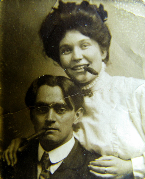 Pale sepia-toned portrait of a mischievous-looking couple. Posed close together with hairstyles and dress that code man and woman, they are leaning against each other with relaxed informality. The man is apparently seated, leaning back against the woman, while she is leaning forward a bit, chin almost resting on his head and her hands on his shoulders. Both are holding cigars in their mouths. The short-haired man is unsmiling, cigar dangling from the corner of his lips, and is dressed in a  dark jacket and tie over a starched-looking white shirt. The woman is smiling, cigar clenched between her teeth, and her hair is styled in a loose updo. She's wearing a high-necked white blouse with short ruffles around the collar and down the front. The blouse is fastened with a brooch at the neck. She has a band-like ring on her left middle finger.
